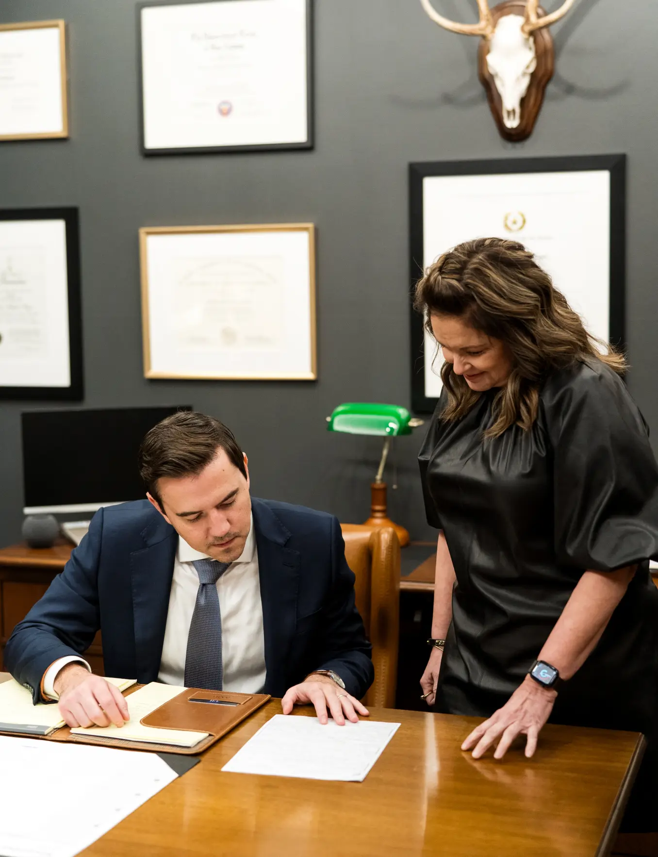 Attorney Austin Hagee writing at his desk while a colleague looks over a document, framed licenses behind